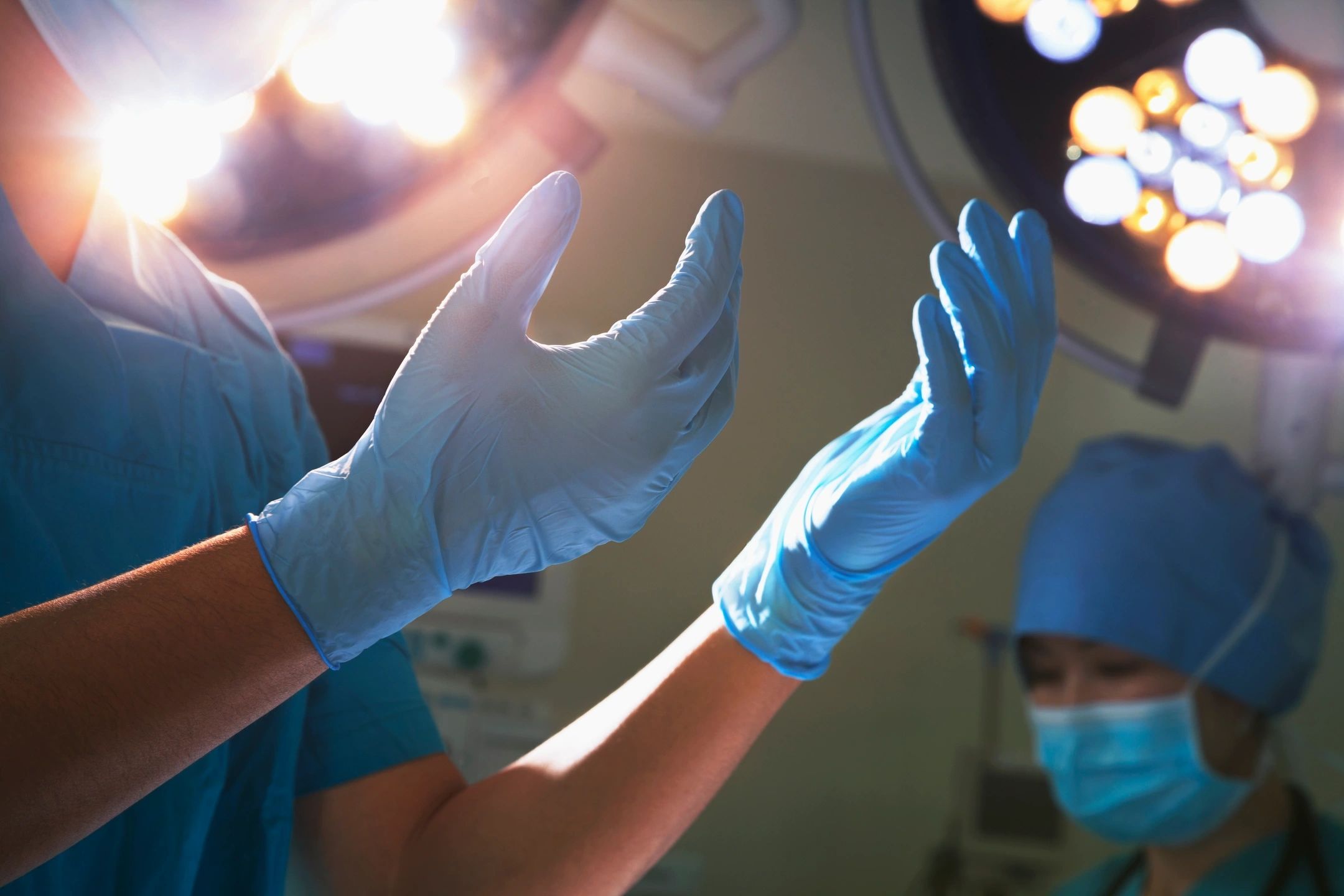 A surgeon in blue scrubs and sterile gloves holding up their hands under bright operating room lights.