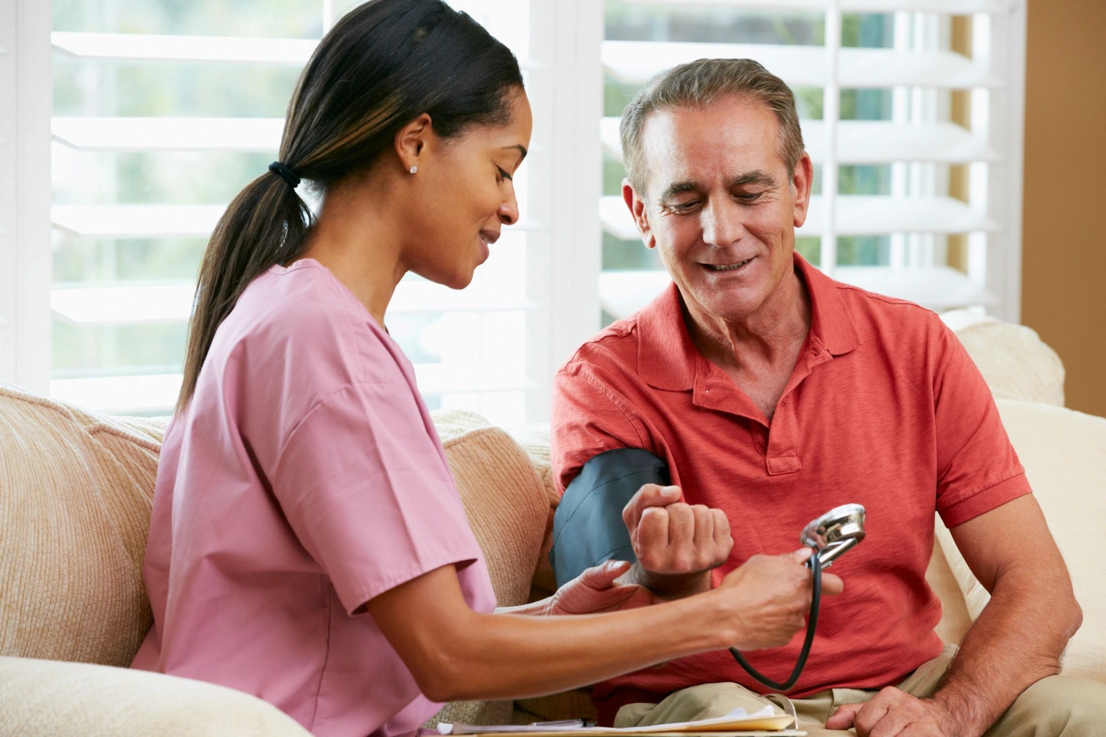 A home health nurse using a manual cuff to check an elderly male patient's blood pressure on a living room couch.