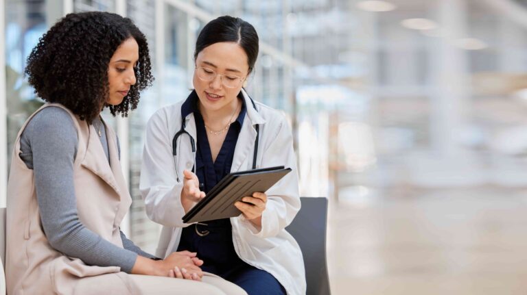 A female doctor wearing a lab coat and glasses explaining test results on a tablet to her patient.